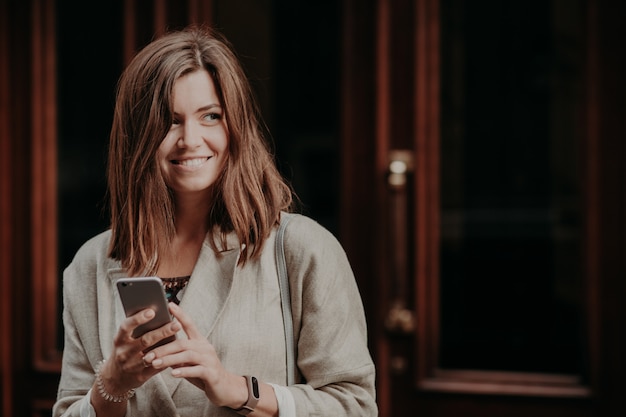 Photo of good looking woman searches information, uses cell phone, dressed in elegant jacket