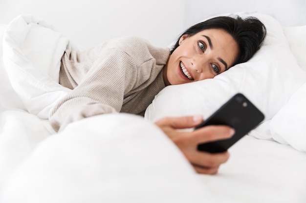 Photo of gorgeous woman 30s using smartphone, while lying in bed with white linen at home