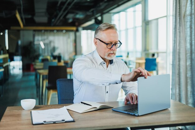 Photo of a grayhaired boss in the office with a laptop a successful and manager man in glasses and a white shirt an investor with a beard sitting on a chair