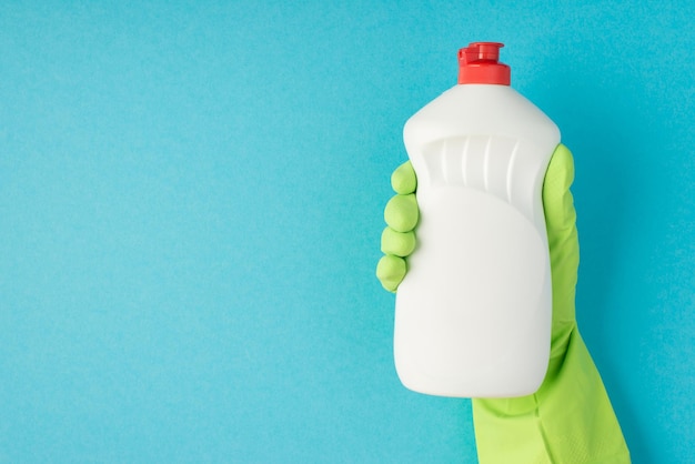 Photo of hand in green rubber glove holding white detergent gel bottle without label on isolated pastel blue background with blank space