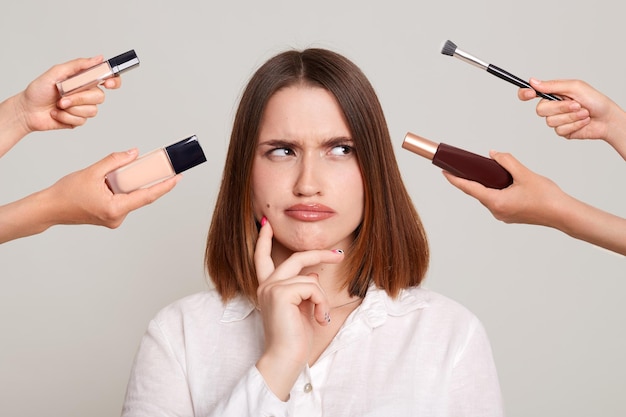 Photo of hands of several beauticians holding their respective equipment giving makeup to attractive woman standing with pensive thoughtful expression