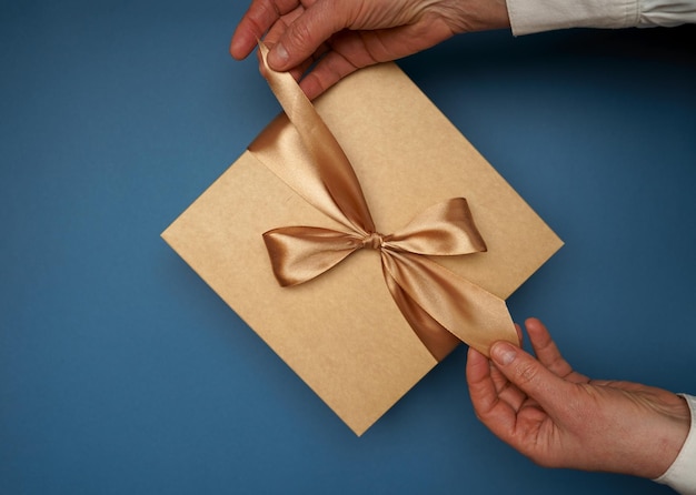 Photo of hands unpacking craft paper gift box with ribbon on isolated blue background