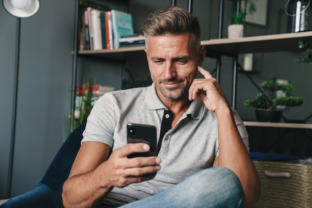 Photo of handsome caucasian man in casual clothing holding smartphone while sitting on armchair at home