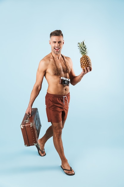 Photo of a handsome excited emotional tourist adult man posing over blue wall holding pineapple and suitcase.