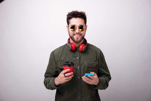 Photo of handsome young guy in sunglasses and headphones, holding a cup and a smartphone