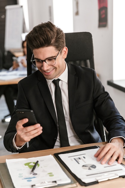 Photo of happy businessman in eyeglasses typing on cellphone and working with documents while sitting at desk in open-plan office