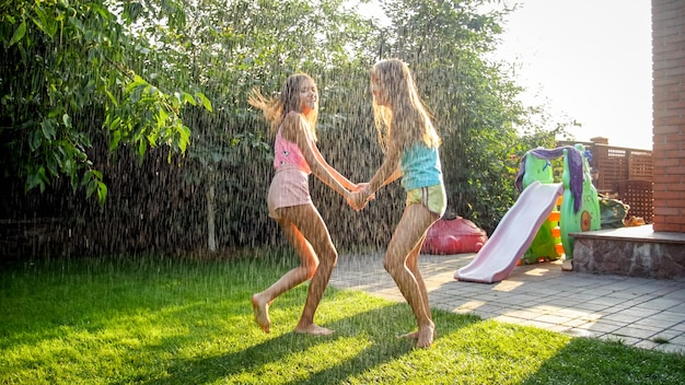 Photo of happy laughing children in wet clothes jumping and dancing under warm rain at garden. Family playing and having fun outdoors at summer