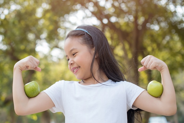 Photo happy Little asian girl child standing with big smile. girl with green apple showing biceps.
fresh healthy green bio nature with abstract blurred foliage and bright summer sunlight