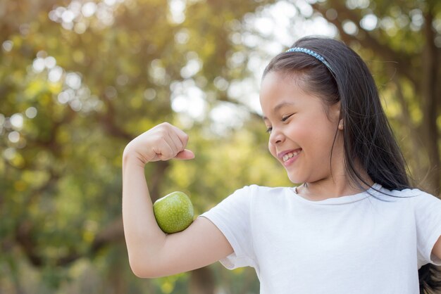 Photo happy Little asian girl child standing with big smile. girl with green apple showing biceps.
fresh healthy green bio nature with abstract blurred foliage and bright summer sunlight