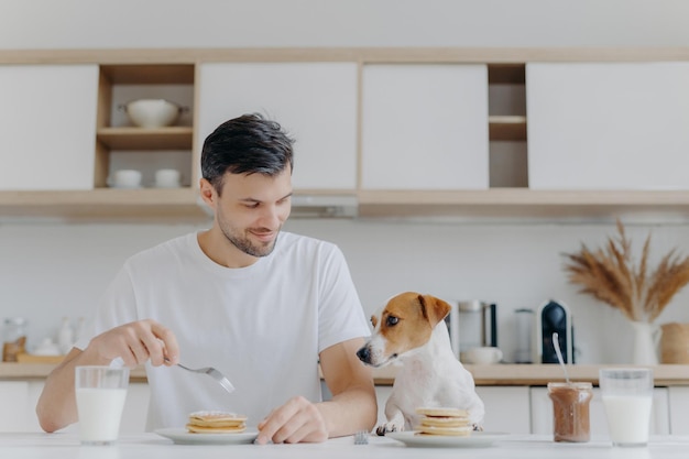 Photo of happy man eats tasty pancakes with fork drinks milk from glass wears white t shirt his jack russell terrier dog poses near has delicious breakfast together with host kitchen background