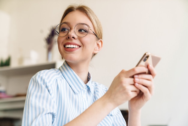 Photo happy nice woman architect in eyeglasses using mobile phone while designing draft at workplace