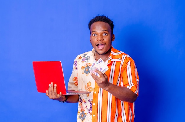 Photo of a happy young afro american handsome man posing isolated over blue wall background using laptop computer holding money