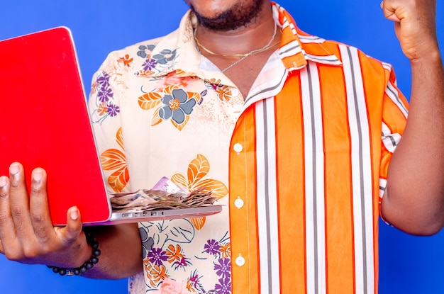 Photo of a happy young afro american handsome man posing isolated over blue wall background using laptop computer holding money