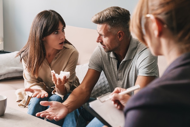 Photo of irritated unhappy couple man and woman having conversation with psychologist on therapy session in room