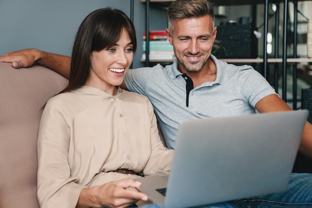 Photo of joyful caucasian couple man and woman smiling while watching laptop on sofa at home