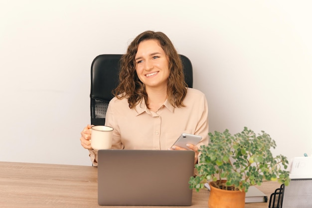 Photo of joyful young office woman in casual sitting at desk and holding cup of coffee and smartphone