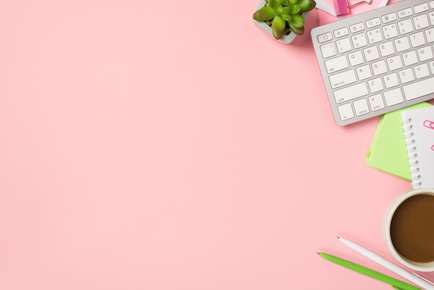 Above photo of keyboard cup of coffee notepad pens paper clips and green plant isolated on the pink backdrop with copyspace