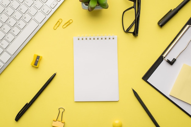 Photo above of keyboard notebook clipboard paperclips pen notes plant sharpener and glasses isolated on the yellow background