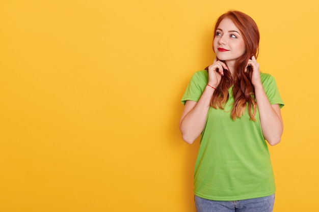 Photo of lovely ginger woman looking aside, looks with interesting expression, has wavy red hair, wearing green casual t shirt, recommends advertisement over yellow wall.