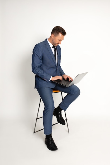 Photo of man 40 years old sits on bar stool on white background in studio. lap top on knees, works, looks into frame, manager posing tiping on laptop