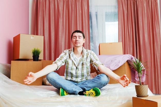 Photo of man sitting in lotus pose on sofa among cardboard boxes