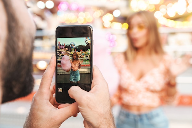 Photo of a man take photo of his shocked emotional woman walking outdoors in amusement park by mobile phone.