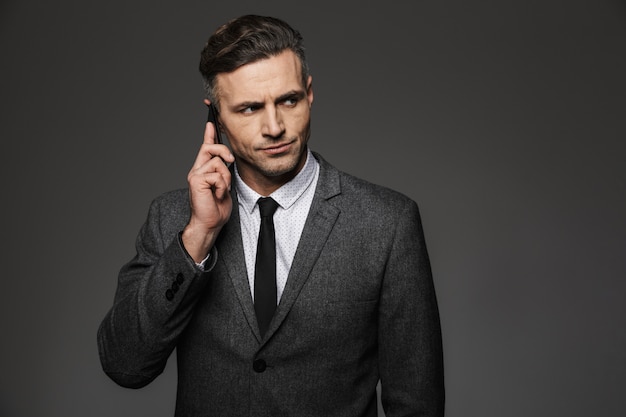 Photo of masculine man 30s dressed in businesslike costume working in office and talking on phone about business, isolated over gray wall