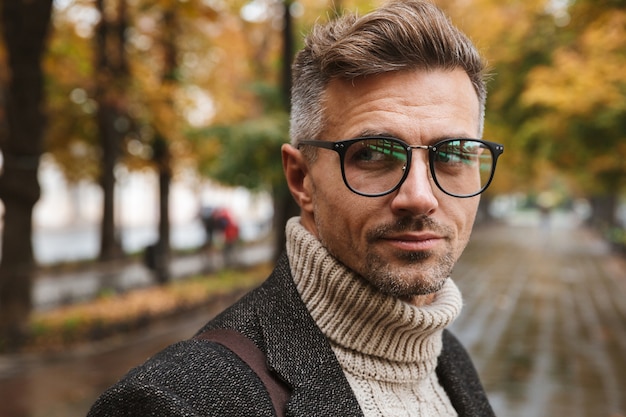 Photo of masculine man 30s wearing eyeglasses, while walking outdoor through autumn park