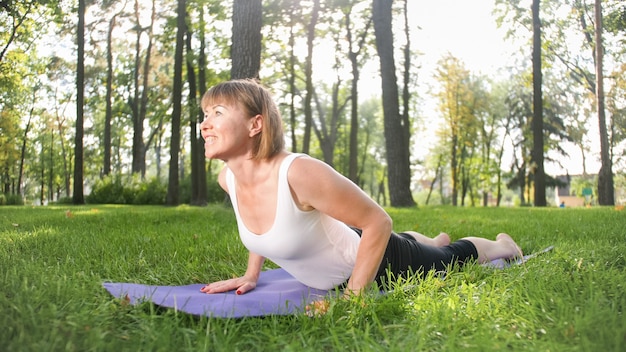 Photo of middle aged woman practicing yoga or fitness on fresh green grass at park. Female physical and mental health. Person in meditation and harmony pf body and soul