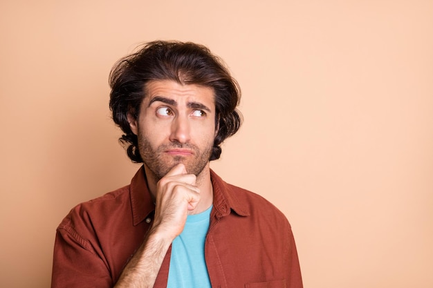 Photo of minded doubtful young guy arm chin look empty space wear brown t-shirt isolated beige color background