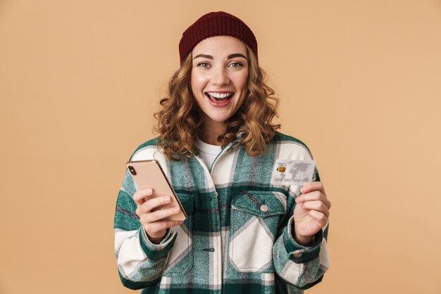 Photo of nice cheerful woman in knit hat holding credit card and cellphone isolated on beige