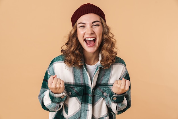 Photo of nice joyful woman in knit hat laughing and making winner gesture isolated on beige