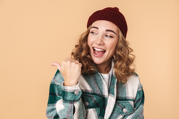 Photo of nice laughing woman in knit hat pointing fingers and looking aside isolated on beige