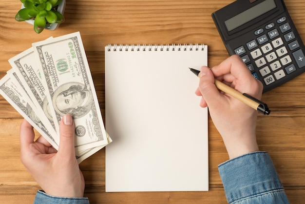 Above photo of notebook pen hands money earnings cash green plant and calculator isolated on the wooden backdrop