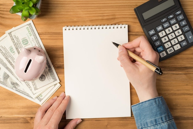 Above photo of notebook pen hands money earnings cash green plant moneybox and calculator isolated on the wooden backdrop