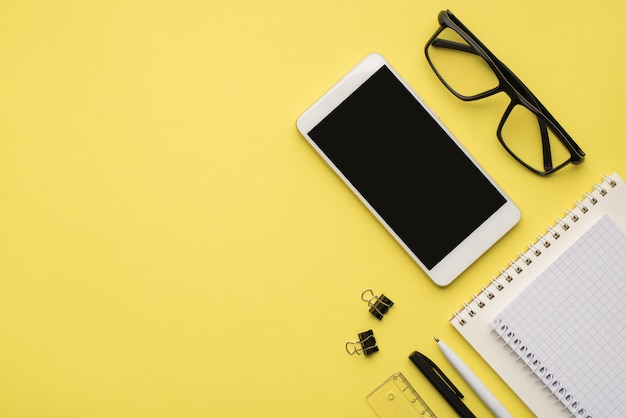 Photo overhead of notebook glasses paperclips phone pen and straightedge isolated on the yellow background