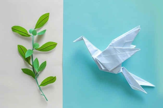 Photo of a paper origami dove holding an olive branch on a light blue background representing peace