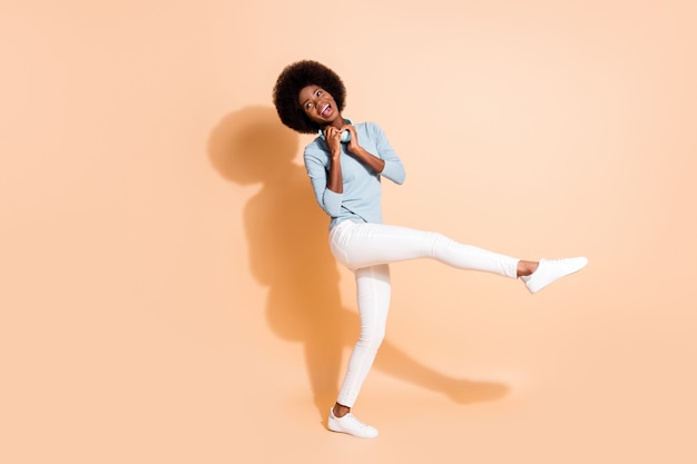 Photo portrait of african american girl holding teal headphones around neck standing on one leg isolated on pastel beige colored background