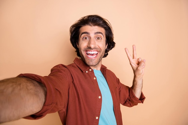 Photo portrait of excited guy showing v-sign taking selfie isolated on pastel beige colored background