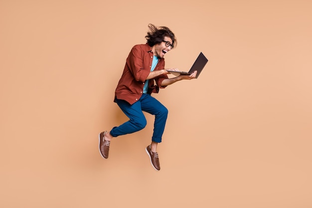 Photo portrait full body view of screaming guy jumping up with laptop in hands isolated on pastel beige colored background