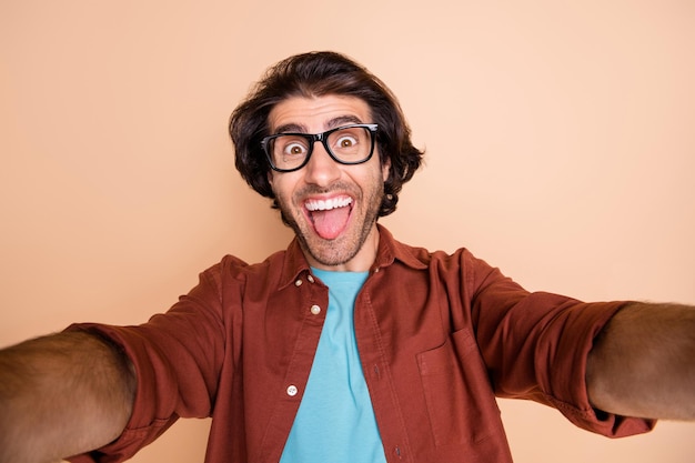 Photo portrait of happy man taking selfie showing tongue isolated on pastel beige colored background