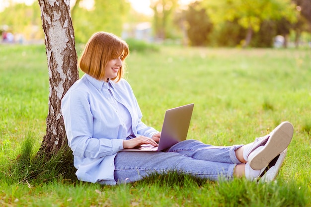 Photo portrait of pretty young woman smiling teeth sitting in green park working on laptop smiling