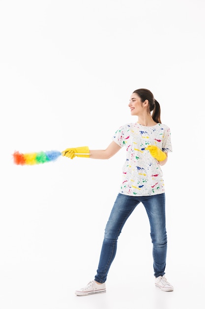 Photo of positive woman 20s wearing yellow rubber gloves for hands protection holding colorful duster while cleaning room isolated over white wall