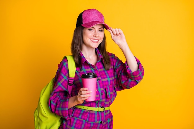 Photo of pretty cheerful teen girl hold cup touch headwear go to school wear pink shirt green belt bag isolated on yellow color background