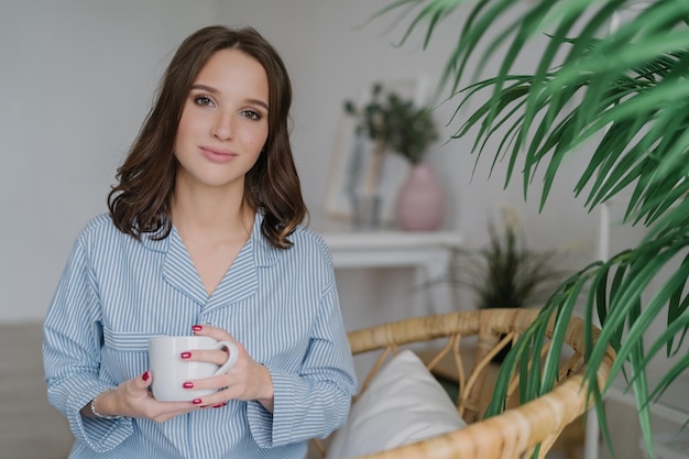 Photo of pretty female with appealing look dressed in casual domestic clothes holds white mug with aromatic coffee or tea sits on wicker furniture indoor with copy space for your advertisement