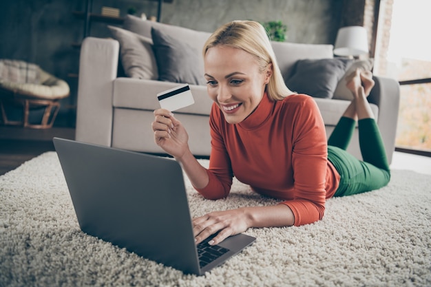 Photo photo of pretty homey lady choosing internet website notebook purchase prepared credit card for payment lying carpet floor near couch casual outfit living room indoors