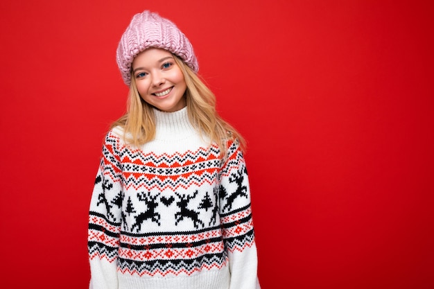 Photo of pretty positive smiling young blonde woman isolated over red background wall wearing winter sweater and pink hat