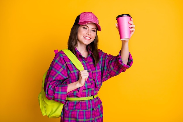 Photo of pretty positive teen girl hold cup greetings wear pink headwear shirt green strap bag isolated on yellow color background