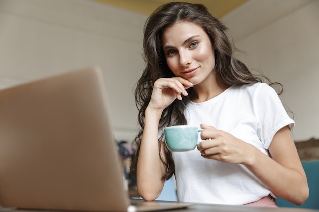 Photo of a pretty positive young woman indoors at home using laptop computer drinking coffee.