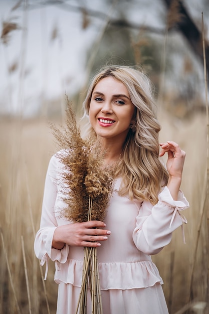 Photo of a pretty smiling girl with long blond curly hair in light long drees standing in a reed field